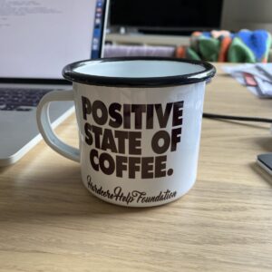 A tin cup on a desk. On the cup, the words “Positive State of Coffee” are written.